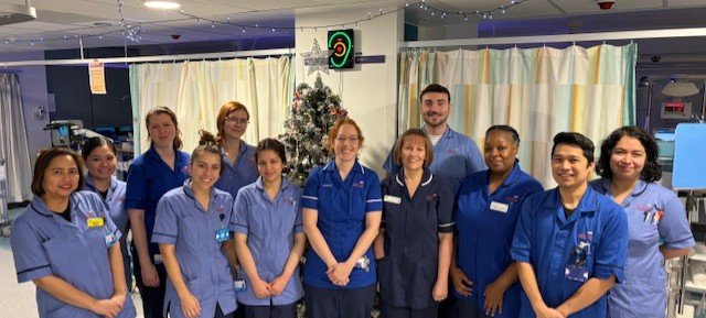 A group of 12 nurses, all in uniform smile at the camera, they are in a hospital ward with a Christmas tree in the background