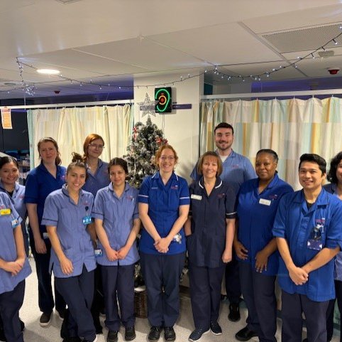 A group of 12 nurses, all in uniform smile at the camera, they are in a hospital ward with a Christmas tree in the background