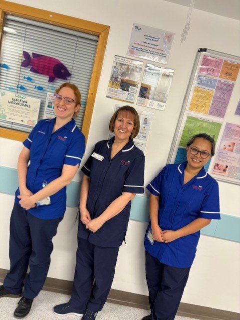 Three nurses in blue uniform smile at the camera in front of a notice board