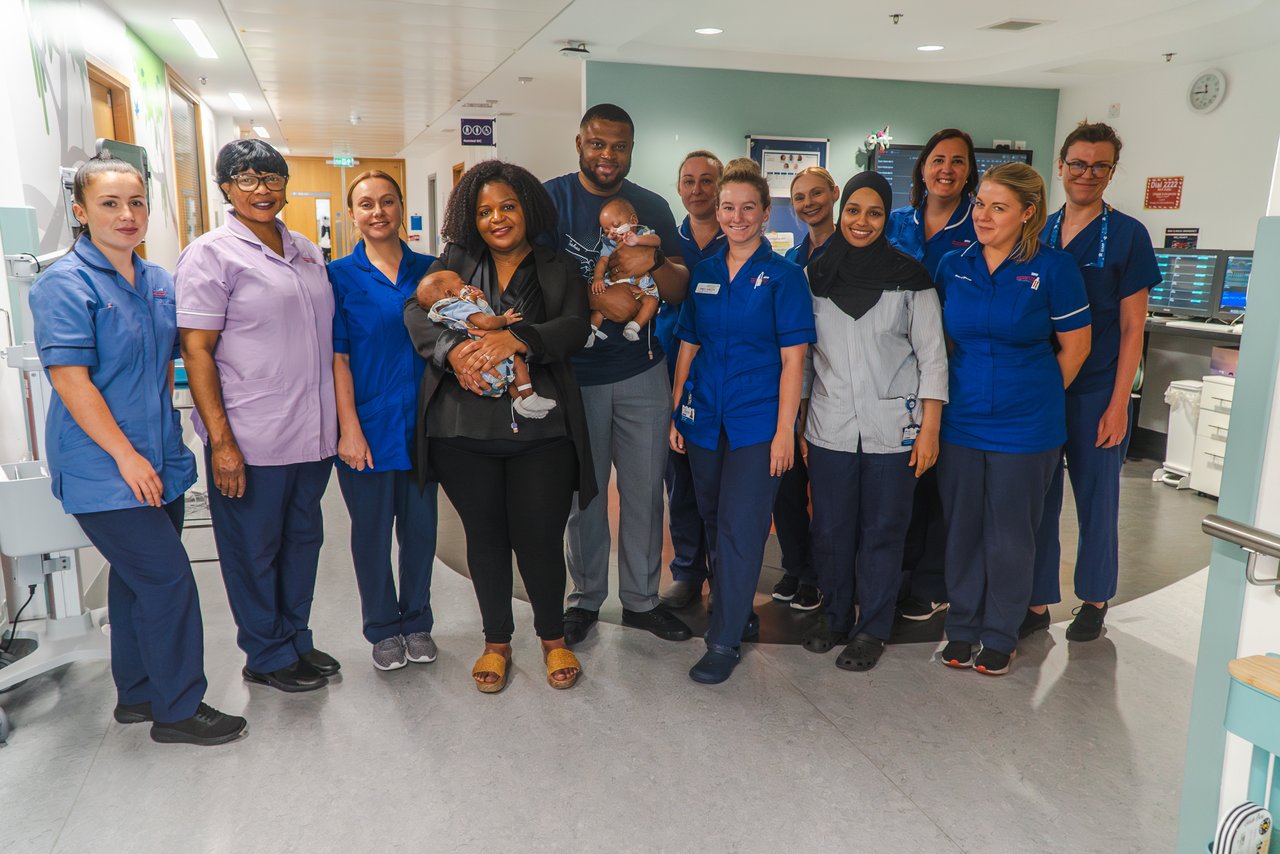 A lady and gentlemen each holding a baby. They are stood with a group of people all wearing blue of purple tops.