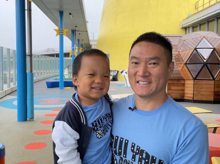 Ollie being held by dad Ricky, both f them are wearing blue t-shirts standing in front of a colourful play area