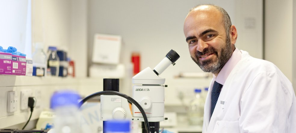 Photo of a man with a beard, wearing a pink shirt, blue tie and white lab coat. He is standing in front of a microscope