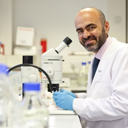 Photo of a man with a beard, wearing a pink shirt, blue tie and white lab coat. He is standing in front of a microscope