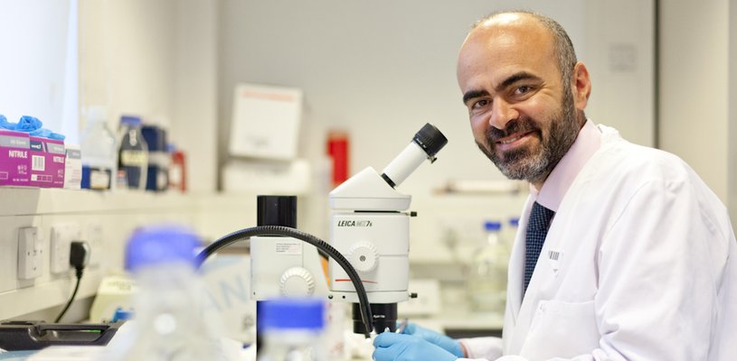 Photo of a man with a beard, wearing a pink shirt, blue tie and white lab coat. He is standing in front of a microscope
