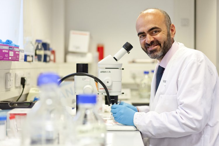 Photo of a man with a beard, wearing a pink shirt, blue tie and white lab coat. He is standing in front of a microscope