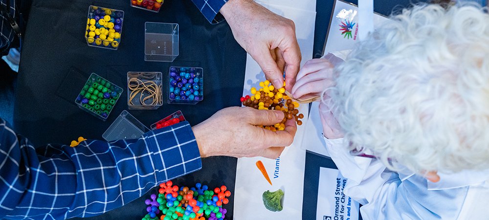 A view from above of adult hands playing with some scientific models and the top of a child's head. The child has bright white hair and is wearing a lab coat. You can't see their faces