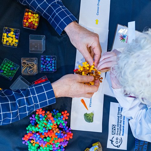 A view from above of adult hands playing with some scientific models and the top of a child's head. The child has bright white hair and is wearing a lab coat. You can't see their faces