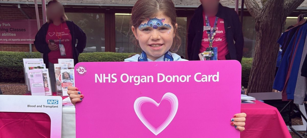 Patient holding a pink NHS blood and transplant card. They are smiling warmly at the camera. Behind them is an NHS Blood and Transplant tent, with two people by an information stand. Their faces are blurred.