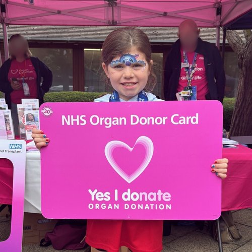Patient holding a pink NHS blood and transplant card. They are smiling warmly at the camera. Behind them is an NHS Blood and Transplant tent, with two people by an information stand. Their faces are blurred.