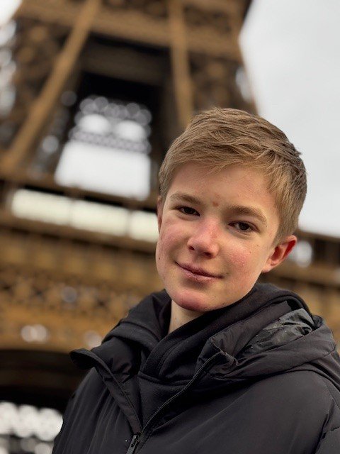 A close up image of a boy in a black coat. He is smiling at the camera. Behind him the Eiffel Tower, it is slightly blurry.
