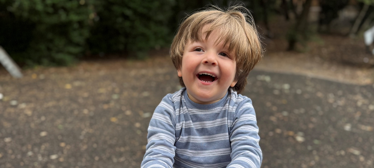 A young boy with brown hair sat on a seesaw. He is smiling and wearing a blue striped top.