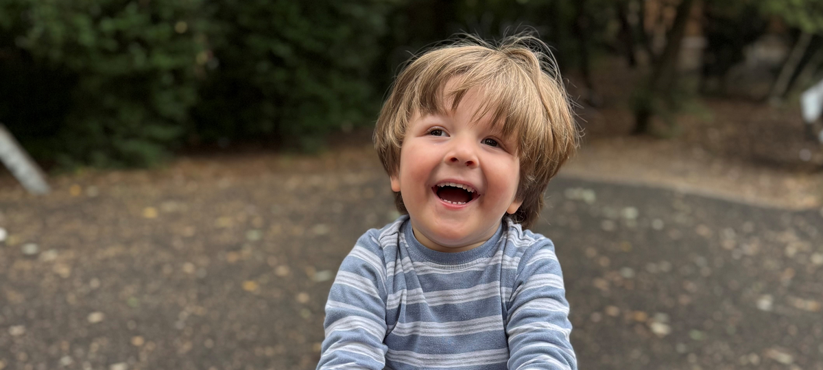 A young boy with brown hair sat on a seesaw. He is smiling and wearing a blue striped top.
