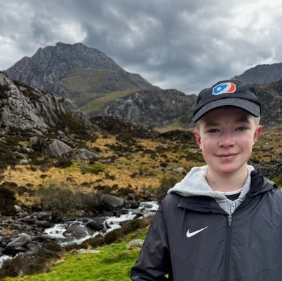A boy stood in front of mountains wearing a black baseball cap and smiling.