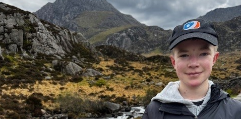 A boy stood in front of mountains wearing a black baseball cap and smiling.