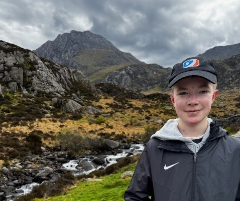 A boy stood in front of mountains wearing a black baseball cap and smiling.