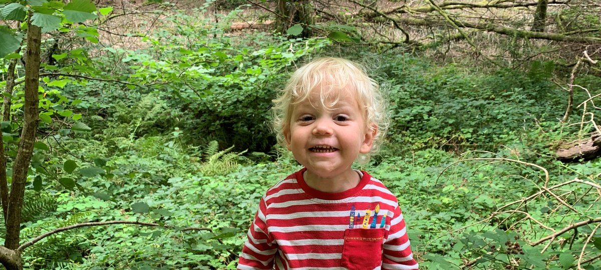 A toddler is sat on a log in a woodland area. They are grinning at the camera and are wearing a stripey top, trousers and socks.