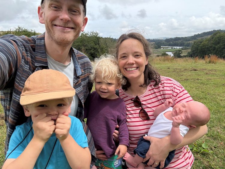 A family are taking a selfie which features a man, women, 2 children and a new born baby. They are all smiling happily at the camera, and the baby is asleep. Behind them is grass and trees.