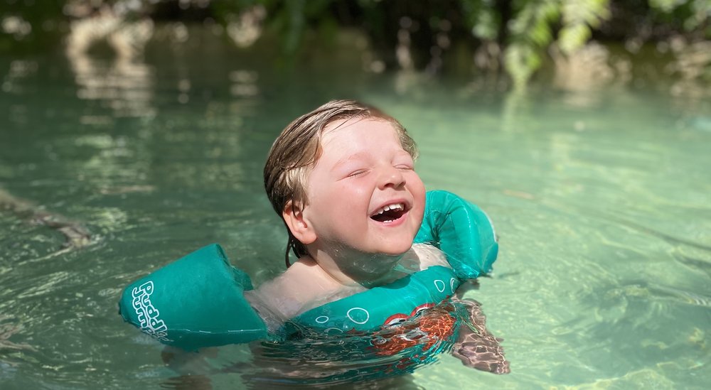 A young boy swimming. He is wearing green arm bands and smiling. His eyes are closed.