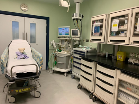 A hospital operating room with a bed holding a teddy bear covered by a blanket. The room includes medical equipment such as monitors, an anaesthesia machine, and storage cabinets with supplies. A clock is mounted above double doors in the background.