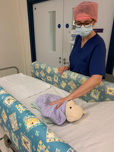 A person in medical scrubs and a hairnet stands beside a hospital bed with colourful padded side rails featuring geometric patterns. A stuffed animal wrapped in a purple blanket lies on the bed.