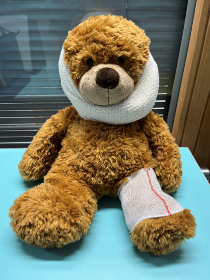 A brown teddy bear sitting on a light blue surface with a bandage wrapped around its head and another around its left leg, which has a red line down the middle. The background features horizontal blinds and part of a wooden frame.