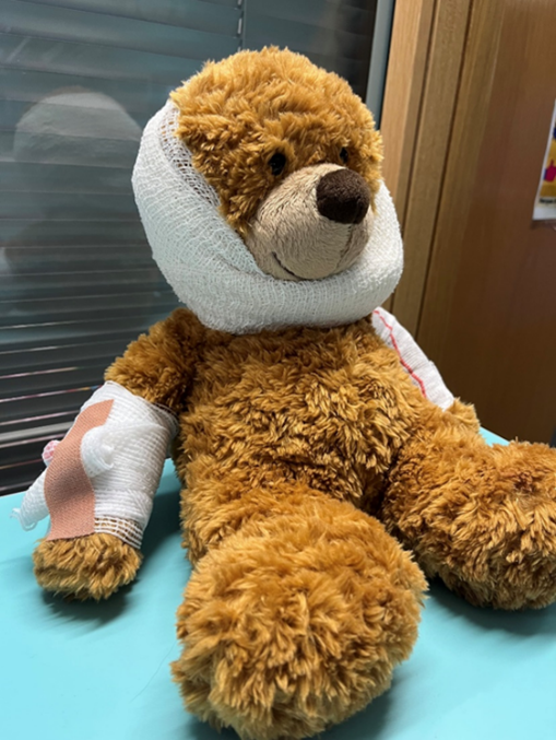 A brown teddy bear sitting on a light blue surface with bandages wrapped around its head and left arm, suggesting it is injured or recovering.
