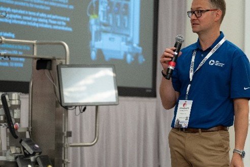 A man holding a microphone is giving a presentation in front of some equipment