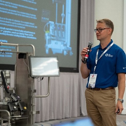 A man holding a microphone is giving a presentation in front of some equipment