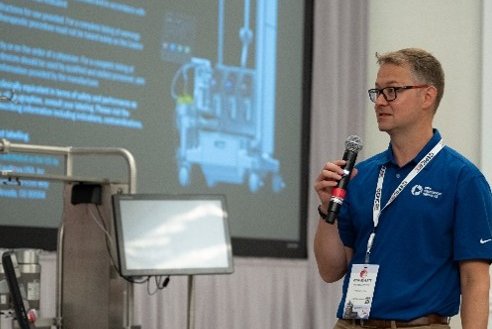 A man holding a microphone is giving a presentation in front of some equipment