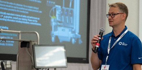 A man holding a microphone is giving a presentation in front of some equipment