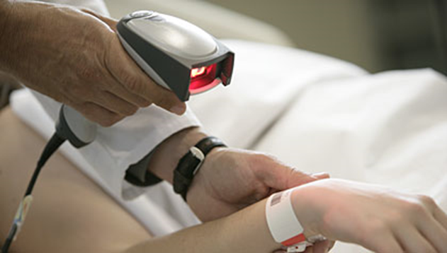 A person in a white coat scans a patient's wristband using a barcode scanner. The patient is lying in bed with their arm extended, and the wristband is white with red markings.