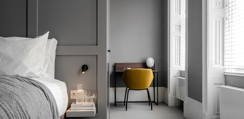 A photo of the bedrooms in the new Powis Place Accommodation, showing a double bed, a bright window, a yellow chair and small desk.