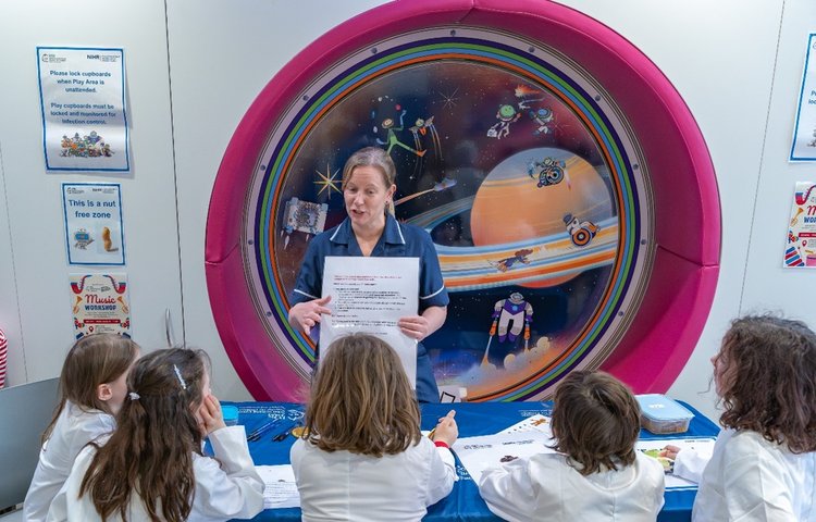 A woman in a navy blue nurses uniform stands in from of a space scene. She is holding up a big piece of paper and 5 children in lab coats are listening to her