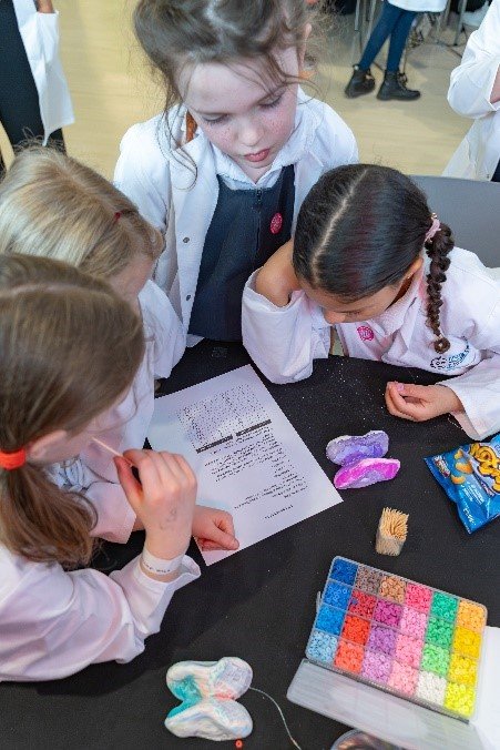 Four girls in lab coats and school unfirm huddle together over a piece of paper