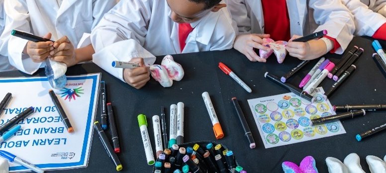 A group of children in lab coats colour in 3-d models - you can see a table and their hands but not their faces