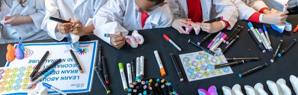 A group of children in lab coats colour in 3-d models - you can see a table and their hands but not their faces