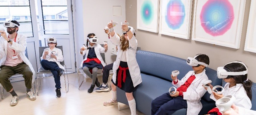 5 children and an adult are in school uniform and lab coats with virtual reality headsets  and controllers. One child is stood up with her hands and face to the sky.
