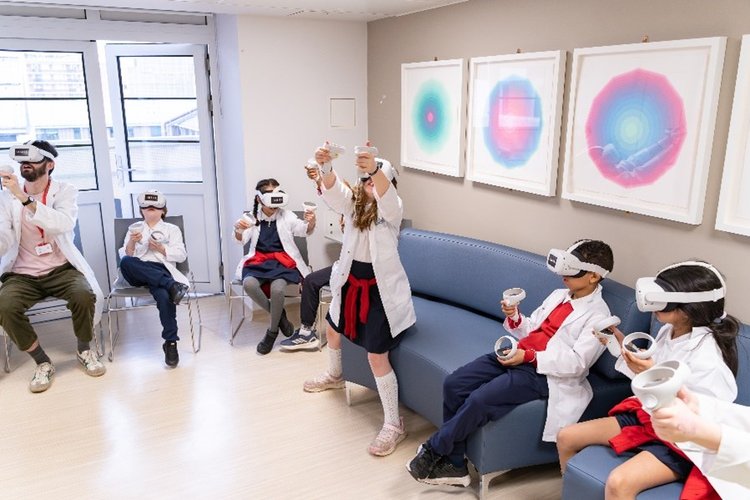 5 children and an adult are in school uniform and lab coats with virtual reality headsets  and controllers. One child is stood up with her hands and face to the sky.