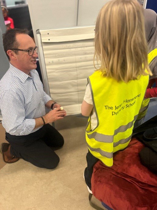 A man kneels down, explaining something to a child with long blonde hair and a high visibility tabard.