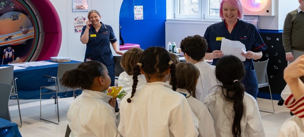 A woman in a navy blue nurses uniform smiles broadly at a group of children in white lab coats