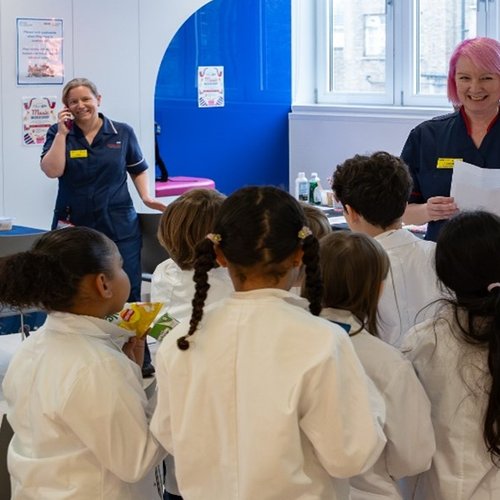 A woman in a navy blue nurses uniform smiles broadly at a group of children in white lab coats