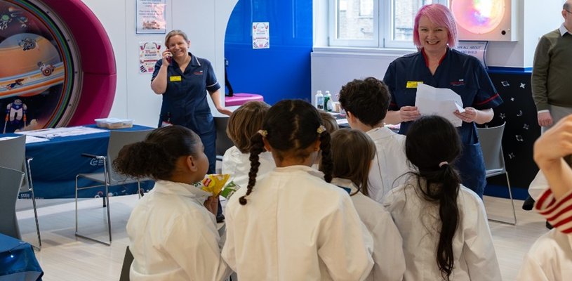 A woman in a navy blue nurses uniform smiles broadly at a group of children in white lab coats