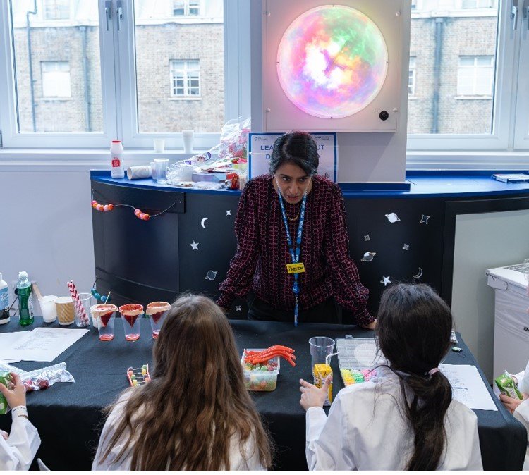 A woman with light brown skin leans on a table covered in beakers and colourful equipment. Two children in lab coats are listening to her