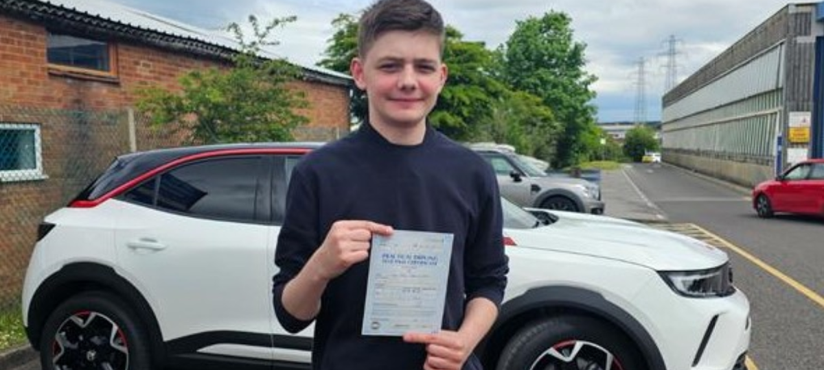 Boy with brown hair stood infront of a white car holding up a piece of paper driving license