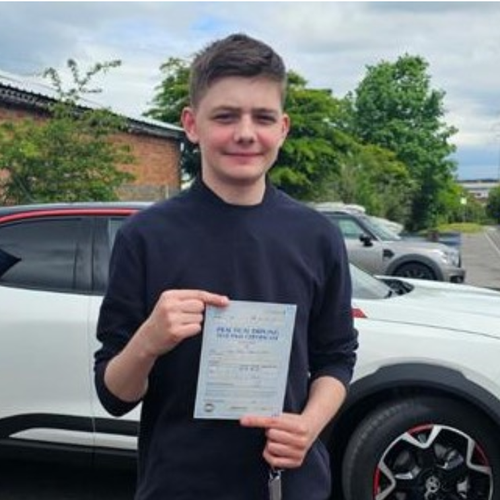 Boy with brown hair stood infront of a white car holding up a piece of paper driving license