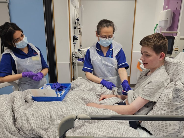 Boy with brown hair sat in a hospital bed while a nurse administers an infusion. Another female nurse is stood smiling.