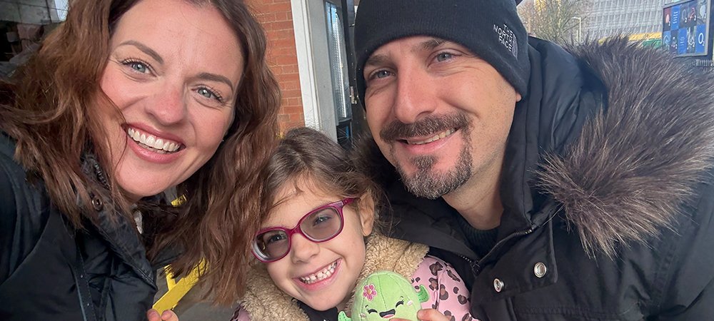 A woman, young girl and a man smile broadly at a the camera in a 'selfie'. The little girl is in the middle in a pink furry coat and glasses. They look really happy.