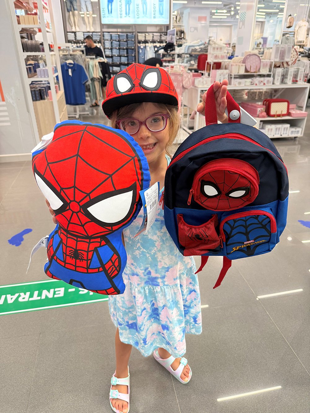 A young girl in a Spiderman hat holds up two spiderman rucksacks and smiles broadly.