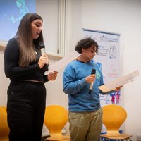 A young man and young woman speaking into a microphone, reading a speech