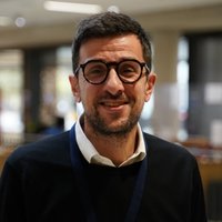 A man in a black jumper and heavy black glasses smiles at the camera